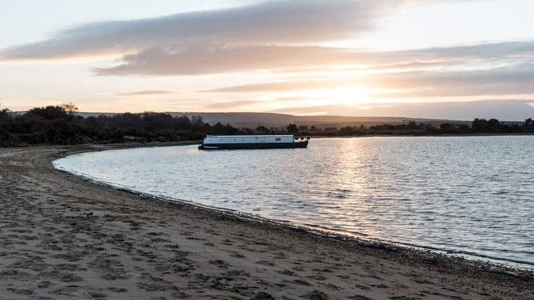 Bay with sandy beach in foreground, and sea beyond lit by sunset. Houseboat in distance.